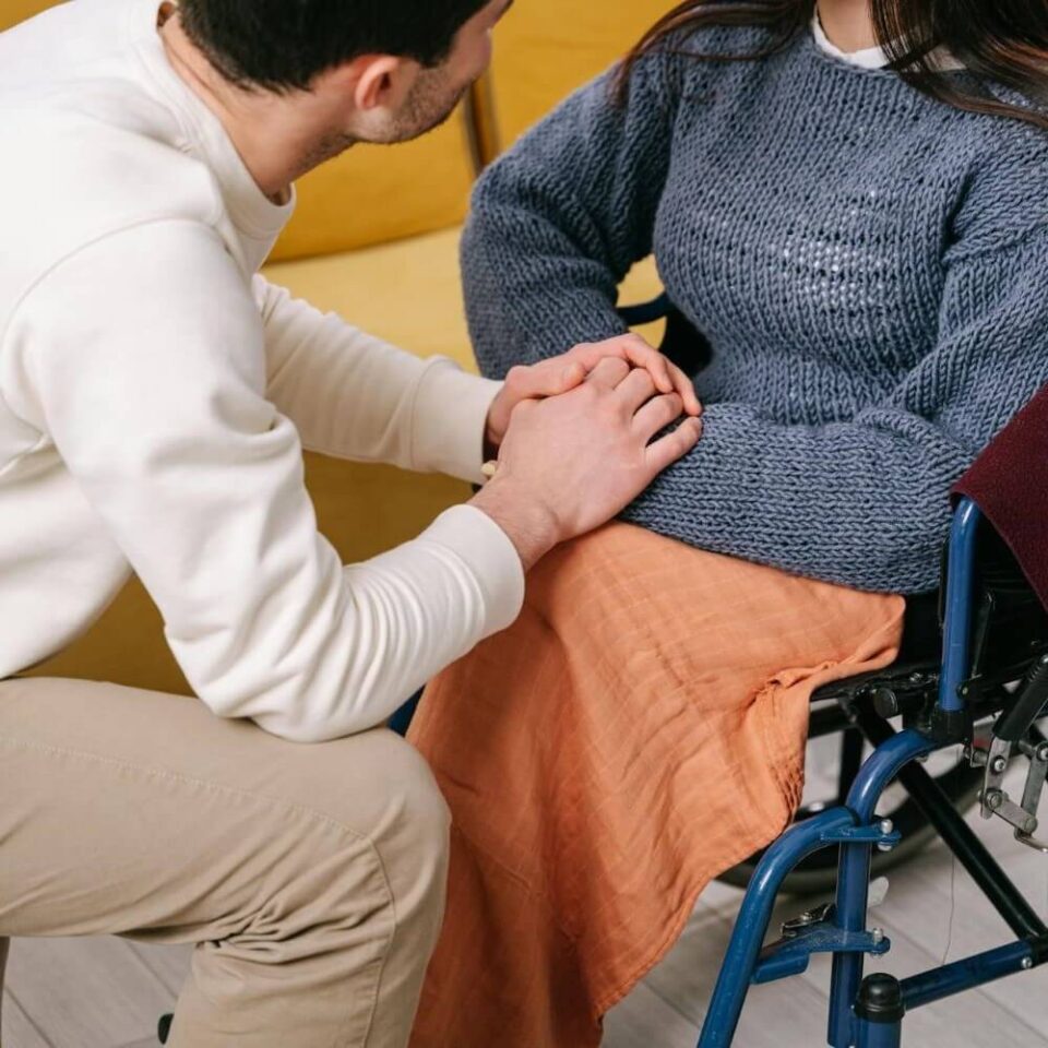 Man crouching beside a woman in a wheelchair, holding her hands, showing compassion and support.