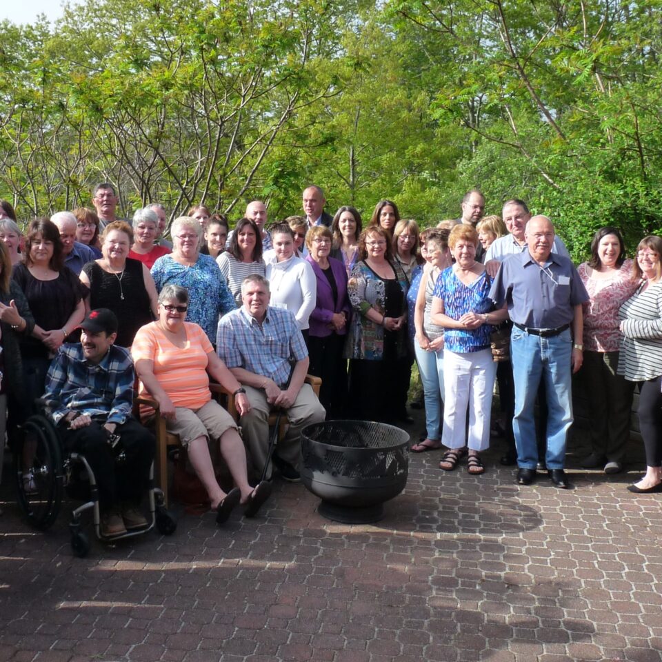 Group of people of assorted age, gender and ability, standing outside, green trees in background.