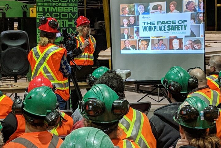 A woman wearing a safety vest and hard hat stands in front of a crew on a shop floor. The slide deck projected behind her reads "The face of workplace tragedy"
