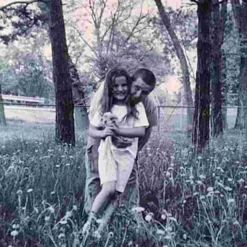 A young girl and her dad embrace in a summer field, with trees behind them. The image is in sepia tone.