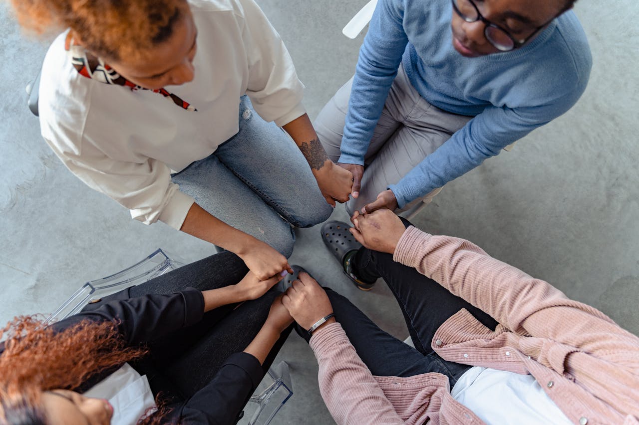 Four people sit close together in a circle, holding hands.