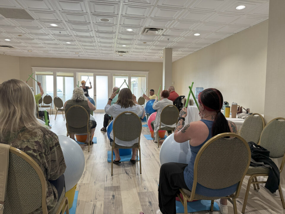 A group of people seated, hold drumsticks in the air as they participate in an activity.