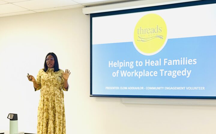 A woman stands beside a screen with a slide displayed beside her. The slide reads "Threads of Life: Helping to Health Families of Workplace Tragedy"