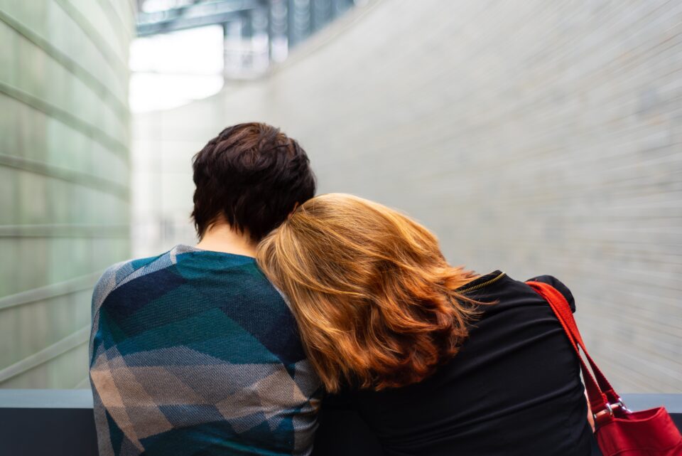 The rear view of two people seated on a bench. One with long, red hair has their head resting on the shoulder of the other person.
