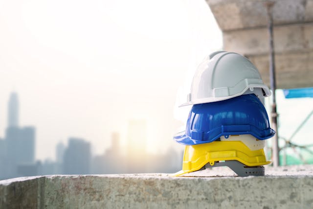 3 hard hats - one white, one blue, and one yellow - are stacked on a concrete ledge.