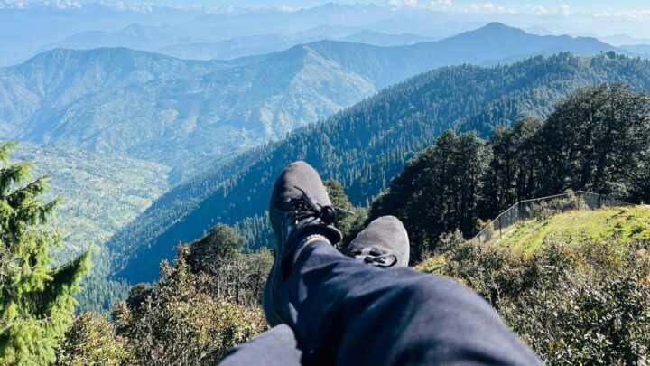 The crossed legs of a person sitting on top of a mountain overlooks several peaks in a mountain valley.